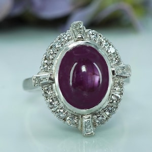 May include: A silver ring with an oval, deep red ruby as the central stone. The ruby is encircled by small, clear, round gemstones. Rectangular gemstones are set on the sides of the ring. The ring is a piece of jewellery.