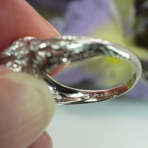 May include: Close-up of a platinum ring with a floral design and numerous small, sparkling stones. The word "PLATINUM" is stamped on the inside of the ring band. The ring is held by a person's fingers.