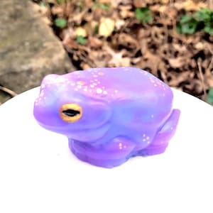 May include: A small purple frog figurine with gold accents and white speckles. The frog has a light blue underside and is sitting on a white surface.