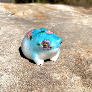 May include: A ceramic frog figurine with a light blue body and white belly. The frog is decorated with pink flower accents and green leaves. The frog is sitting on a stone surface.