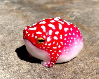 Puede incluir: Figura de rana artesanal. La rana tiene un cuerpo rojo brillante con manchas blancas, que se transforma en rosa y blanco en el vientre. Tiene ojos dorados y está colocada sobre una superficie texturizada, destacando su diseño único.