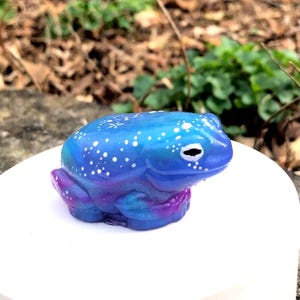 May include: A small, blue and purple frog figurine with white speckles. The frog is sitting on a white surface.