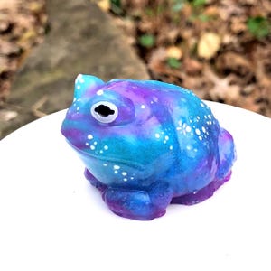 May include: A whimsical frog figurine in shades of blue, purple, and teal, speckled with white dots. The frog has a large, expressive eye and is posed on a white surface. The figurine is a decorative item.