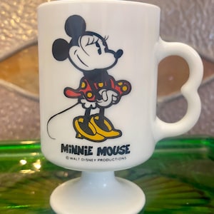 Op de afbeelding: Witte keramische mok met een cartoon afbeelding van Minnie Mouse in een rode jurk met gele stippen. De mok heeft een hartvormig handvat en een voet. De tekst "Minnie Mouse" en "Walt Disney Productions" staat op de mok gedrukt.