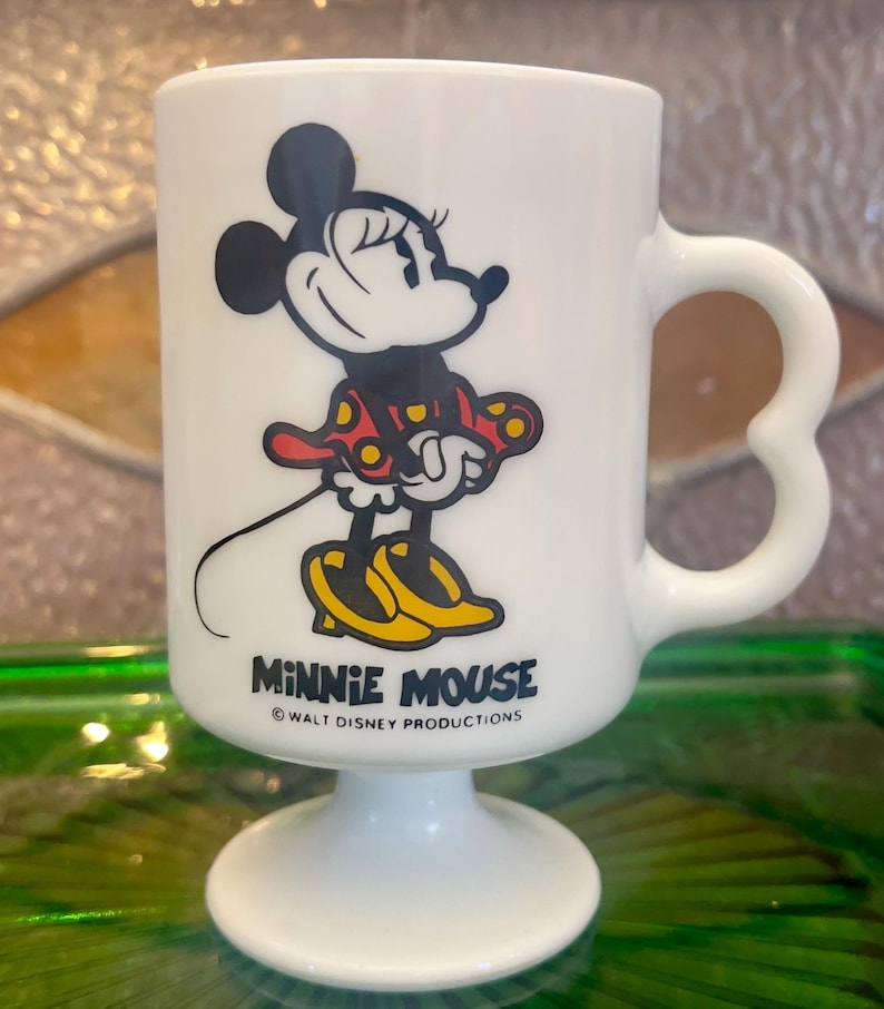 Op de afbeelding: Witte keramische mok met een cartoon afbeelding van Minnie Mouse in een rode jurk met gele stippen. De mok heeft een hartvormig handvat en een voet. De tekst "Minnie Mouse" en "Walt Disney Productions" staat op de mok gedrukt.