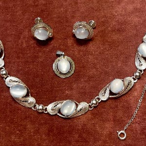 May include: A vintage jewelry set featuring a bracelet, pendant, and earrings. The silver-toned pieces have an openwork design and feature oval, milky-white cabochon stones. The bracelet is connected with a chain.