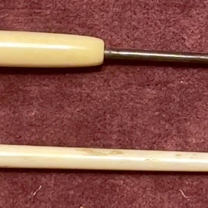 May include: Two antique ivory-colored tools are displayed against a burgundy background. One tool has a curved metal hook and a rounded handle. The other is a long, slender, pointed tool with decorative detailing.