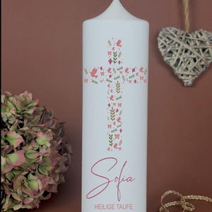 Könnte beinhalten: Weiße Stumpenkerze mit einem Kreuzdesign aus rosa Vögeln, Schleifen und Blättern. Der Name "Sofia" steht in rosa Schrift unter dem Kreuz, darunter "HEILIGE TAUFE" und das Datum "02. November 2025". Die Kerze steht auf einem Holzsockel.