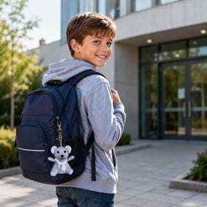 Könnte beinhalten: Ein marineblauer Rucksack mit einem kleinen, hellgrauen Plüschbären-Schlüsselanhänger am vorderen Reißverschluss. Der Bär hat einen weißen Schal, schwarze Augen und eine schwarze Nase und hängt an einem silberfarbenen Karabinerhaken.