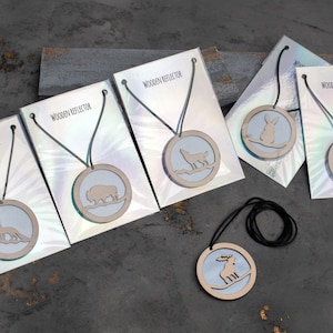 Op de afbeelding: Vijf houten kettingen met verschillende dieren silhouetten op een grijze achtergrond. Elke ketting heeft een zwart koord en een ronde houten hanger met een ander dier uitsparing. De dieren zijn een vos, een bizon, een wolf, een konijn en een hert. De tekst "Wooden Reflector" is op elke kaart gedrukt.
