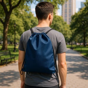 May include: A navy blue drawstring backpack with black drawstrings is worn over the shoulders. The backpack has a textured surface and is being worn in a park setting. The person is wearing a gray t-shirt.