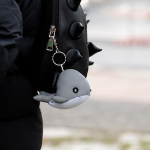 Peut inclure: Un porte-clés en peluche en forme de baleine grise et blanche attaché à un anneau en métal argenté. La baleine présente un petit œil bouton noir avec un détail en croix blanche. Le porte-clés est fixé à la tirette d'un sac à dos noir texturé à pointes.