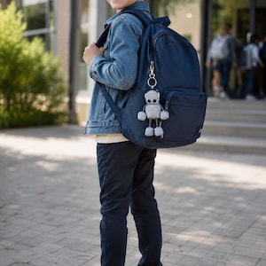 Könnte beinhalten: Ein marineblauer Rucksack mit einem silberfarbenen Roboter-Schlüsselanhänger am Reißverschluss. Der Rucksack verfügt über ein Hauptfach und eine Vordertasche sowie verstellbare Schultergurte und eine robuste Konstruktion für den täglichen Gebrauch.