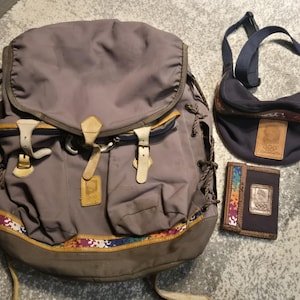 May include: A selection of travel bags and accessories. Includes a large grey backpack with leather accents, a navy blue crossbody bag, and a small wallet. All items feature a colourful trim and a logo.