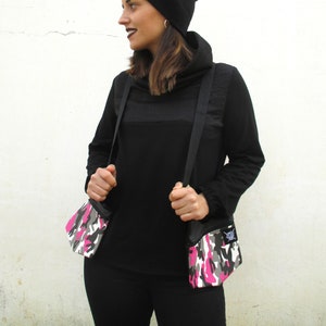 Puede incluir: Una persona lleva una camisa negra de cuello alto con una boina negra y dos bolsos de bandolera de camuflaje con detalles rosas.
