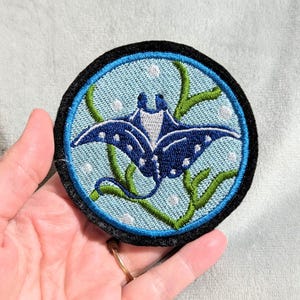 May include: A circular embroidered patch featuring a dark blue manta ray swimming in light blue water. The design includes green seaweed, white bubbles, and a thick black border. The embroidery is detailed with white spots on the ray's wings.