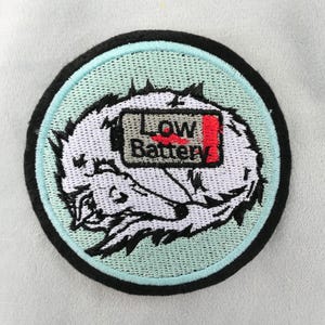 May include: Embroidered patch with a white wolf design inside a light blue circle, with a black border. The patch features the text "LOW BATTERY" and a battery icon with a red indicator.