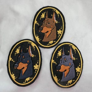 May include: Three embroidered patches featuring Doberman Pinscher dog portraits. Each patch has a black oval border with gold floral accents and a gold stitched edge. The dogs are depicted in shades of brown, blue, and gray.