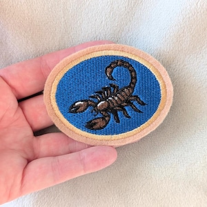 May include: An oval-shaped embroidered patch featuring a brown scorpion centered on a bright blue background. The patch is framed by a thin gold-colored border and a wider, light tan outer edge, creating a distinct layered appearance.
