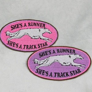 May include: Two embroidered patches with an oval shape. The patches feature a running dog illustration in white, set against a pink and purple background. Text on the patches reads "SHE'S A RUNNER" and "SHE'S A TRACK STAR".
