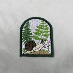 May include: Embroidered patch featuring a snail, ferns, and small pink flowers. The snail has a brown shell and a cream-colored body. The patch is framed by a dark green border, creating a nature-themed design.