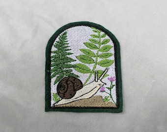 May include: Embroidered patch featuring a snail, ferns, and small pink flowers. The snail has a brown shell and a cream-colored body. The patch is framed by a dark green border, creating a nature-themed design.