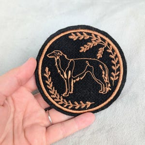 May include: A circular embroidered patch featuring a black background with a copper-colored outline of a Borzoi dog standing in profile. The dog is framed by a delicate, circular wreath of copper-colored leaves and branches stitched onto the fabric.