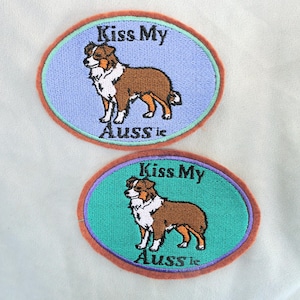 May include: Two oval embroidered patches featuring a brown and white Australian Shepherd dog. The top patch has a light blue background, and the bottom patch has a teal background. Both patches display the black text Kiss My Aussie.