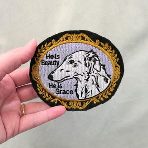 May include: Embroidered patch featuring a Borzoi dog portrait within an oval frame. The frame has a gold floral design on a black background. Text on the patch reads "He Is Beauty, He Is Grace."