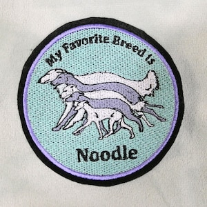 May include: Embroidered patch with a light blue background, featuring a stack of greyhound dog silhouettes in white and lavender. The text "My Favorite Breed Is Noodle" is embroidered around the edge in black and white.