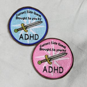 May include: Two embroidered patches with the text "Current Side Quest Brought to you by ADHD". One patch is blue with a sword and compass design, the other is pink. Both have a brown border.