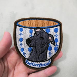 May include: Embroidered patch featuring a black greyhound dog inside a teacup design. The teacup has a brown rim and base, with blue floral patterns. The text "Earl Greyhound" is at the bottom of the patch. The patch is held in a hand.