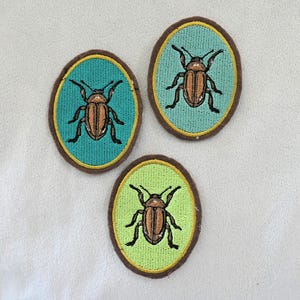 May include: Three embroidered oval patches featuring a brown beetle design. Each patch has a different colored background: teal, light green, and lime green. The patches are outlined with a brown border.