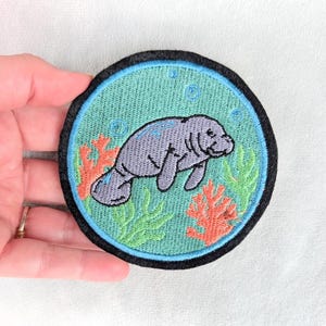 May include: A circular embroidered patch featuring a grey manatee swimming in a light blue sea. The design includes orange coral, green seaweed, and small blue bubbles, all enclosed within a thick black border stitched onto the fabric edge.