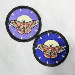 May include: Two embroidered patches featuring a moth design. The moth is brown, orange, and red, set against a yellow sun and a purple or blue background. Each patch has a black border.