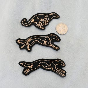 May include: Three black embroidered patches in the shape of running greyhounds with tan thread detailing. A quarter coin is next to the patches for size comparison. The patches are on a light blue fabric.