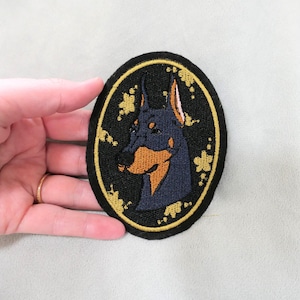 May include: Oval-shaped embroidered patch featuring a Doberman Pinscher dog. The dog's head is depicted in navy blue and tan against a black background, framed by a gold border with floral accents. A unique accessory for dog lovers.