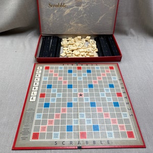 May include: A vintage Scrabble board game set. The red-trimmed game board is open, displaying a grid of squares with point values. The set includes letter tiles and black tile racks, all inside a red box with the word "Scrabble" on the lid.