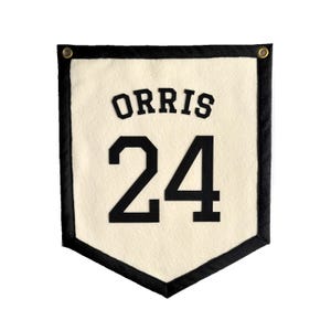 May include: A cream-colored felt pennant with a black border and two brass grommets at the top corners. The word ORRIS is printed in black block letters above the large black number 24, which is centered on the fabric surface.