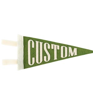 May include: A triangular green felt pennant flag featuring the word CUSTOM printed in large, off-white capital letters across the center. The left side of the pennant includes two short, off-white fabric ties extending horizontally from the base.