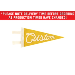 Puede incluir: Un banderín triangular de fieltro color amarillo mostaza con la palabra Custom en letras cursivas blancas. El banderín tiene una franja vertical blanca en el lado izquierdo con dos lazos de tela blanca. El texto superior dice: *PLEASE NOTE DELIVERY TIME BEFORE ORDERING AS PRODUCTION TIMES HAVE CHANGED!