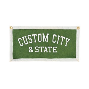 Puede incluir: Un estandarte rectangular de fieltro verde con un borde blanco y dos ojales de latón en las esquinas superiores. El texto CUSTOM CITY & STATE está impreso en letras mayúsculas blancas, con la primera línea arqueada en fuente universitaria.