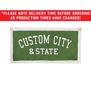 May include: A rectangular green felt wall banner featuring white block lettering that reads CUSTOM CITY & STATE. The banner has a white border and two brass-colored metal grommets at the top corners for hanging purposes on a wall or display.