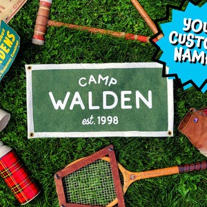 May include: A rectangular green felt pennant with white text reading CAMP WALDEN est. 1998, featuring metal grommets in each corner. The banner is surrounded by a croquet mallet, a tennis racket, a plaid thermos, and a brown leather boot.