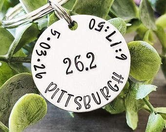 May include: A round, silver-colored keychain tag with the text "26.2 PITTSBURGH" in black. The tag also includes the date "5.03.26" and the time "05:17:50". The tag is attached to a silver keyring and is surrounded by green leaves.