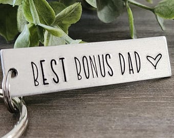 May include: A silver rectangular keychain tag with the words "BEST BONUS DAD" in black lettering and a small heart symbol. The keychain is attached to a silver split ring. Green leaves are in the background.
