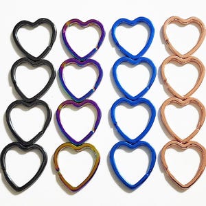 May include: An assortment of heart-shaped metal clasps in various colors. The clasps are arranged in rows and columns, with colors including black, iridescent, blue, and rose gold. These clasps are likely used for jewelry making or crafting projects.