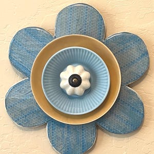 May include: A decorative flower made of ceramic plates. The flower has six blue petals with a dotted pattern. The center features a yellow plate, a light blue plate, and a white knob with a black center.