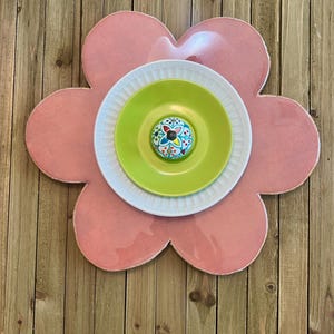 May include: A decorative flower made of a pink, petal-shaped base with a white plate, a green plate, and a colorful knob in the center. The flower is mounted on a wooden background.