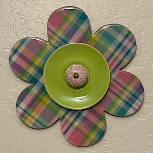 May include: A green and pink plaid flower-shaped wall decor with a green ceramic plate in the center. The plate has a pink ceramic knob in the center.